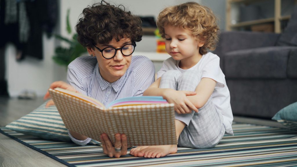 Mother and young son lying on carpet, engaged in reading a book together happily.